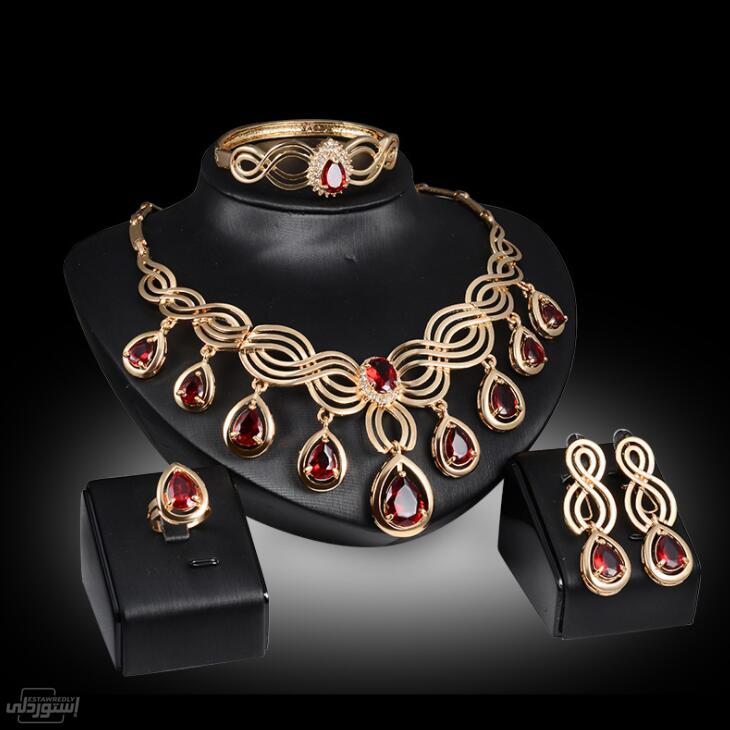 طقم مجوهرات عقد وأسوة وخاتم وحلق ذات تصميمات جذابة ذات لون ذهبي بها دلايات من حجارة حمراء اللون 